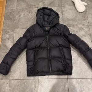 Svart pufferjacka från Colmar - Säljer en svart pufferjacka från Colmar med huva och två snedställda fickor med dragkedja. Jackan har ett stilrent, quiltat mönster och Colmar-logga på ärmen. Perfekt för kalla dagar och har en modern, loose passform. Har haft den några veckor efter jag köpte den av förra ägaren, Märket där bak har tappat lite färg men är jättefin. Säljer snabb affär 2000