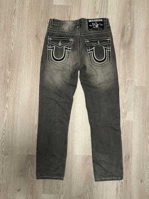 True Religion jeans grå W34  - Svarta jeans från True Religion, modell Joey Super T. Snygga kontrastsömmar i vitt och ikoniska bakfickor med lock och broderad logga. Klassisk femficksdesign, rak passform och slitningar för en cool look. Perfekt för dig som gillar streetwear och detaljer. Den saknar knapp på backfickan  Längd:112 cm Midja:42 Raise:27 Lår:27