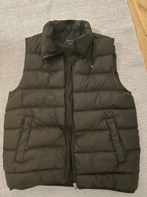 Svart dunväst från GANT - Säljer en svart, quiltad dunväst från GANT med hög krage och två snedställda fickor framtill. Västen har dragkedja hela vägen och klassisk GANT-logga på bröstet. Perfekt för lager-på-lager och snygg till både hoodie och t-shirt.
