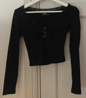 Svart långärmad topp från Cider - Snygg svart långärmad topp från Cider med v-ringad hals och spetsdetalj längs kanten. Toppen har fyra knappar framtill och är croppad i modellen. Tajt tröja samt en kortare modell. Perfekt för dig som gillar stilrena och trendiga plagg. (Rekommenderar som pyjamaströjan)