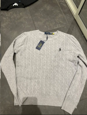 Grå kabelstickad tröja Polo Ralph Lauren - Snygg grå kabelstickad tröja från Polo Ralph Lauren med klassisk rund hals och diskret broderad logga på bröstet. Tröjan har långärmad passform och är tillverkad i mjuk bomull, perfekt för dig som gillar stilrena och tidlösa plagg.