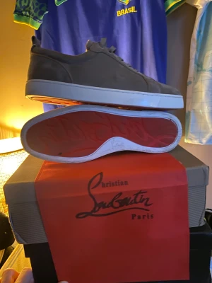 Christian Louboutin grå mocka sneakers - Snygga grå sneakers från Christian Louboutin med klassisk röd sula och diskret logga. Skorna har rund tå, platt vit sula och är tillverkade i mjuk mocka med snörning. Perfekta för dig som vill ha en lyxig och stilren look.