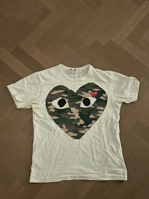 Vit Comme des Garçons Play t-shirt - Vit t-shirt från Comme des Garçons Play med stort hjärta i camouflage-mönster och ögon på bröstet. Liten röd hjärtlogga på vänster sida. Klassisk rund hals och korta ärmar. Perfekt statement-plagg för dig som gillar streetstyle.