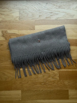 Coffee färgad halsduk från Polo Ralph Lauren - Snygg coffee färgad halsduk från Polo Ralph Lauren med klassisk broderad logga i brunt och blått. Halsduken är rektangulär med långa fransar i kanterna och är tillverkad i mjuk ull. Perfekt för att ge din outfit en clean och stilren touch.