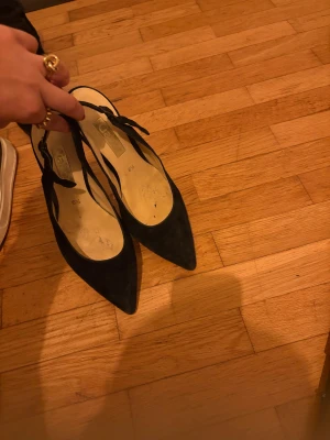 Svarta mocka pumps från Gabor - Snygga svarta pumps från Gabor i mjuk mocka med spetsig tå och låg klack. Skorna har en elegant rem med spänne runt hälen och en stilren, klassisk form som ger en chic vibe. Perfekta för dig som gillar tidlös design och vill ha något som sticker ut.