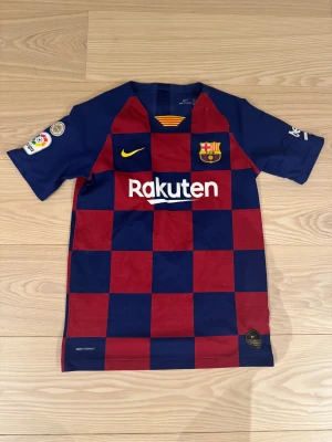 FC Barcelona Messi #10 matchtröja - Säljer en FC Barcelona matchtröja från Nike med Messi #10 på ryggen. Tröjan har rutig design i mörkblått och vinrött, korta ärmar och gula detaljer som klubbmärke, Nike-logga och nummer. Materialet är lätt och ventilerande polyester, perfekt för fotboll.