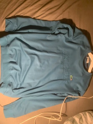 Blå sweatshirt från Lacoste i M passar även s och xs - Snygg blå sweatshirt från Lacoste med broderad logga och krokodilemblem på bröstet. Tröjan har rund hals, ribbade muddar och lång ärm. Perfekt för dig som gillar klassisk streetwear med en sportig touch.