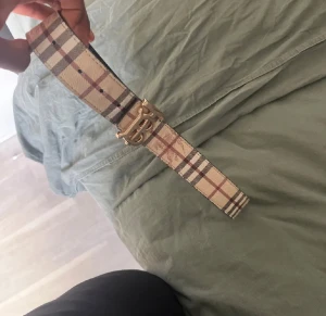 Burberry rutig skinnbälte med guldspänne - Säljer ett snyggt Burberry bälte i skinn med klassiskt rutigt mönster i beige, brun och svart. Bältet har ett stilrent guldspänne med Burberrys ikoniska 'B'-logga framtill. Perfekt för att lyfta din outfit med en lyxig touch.