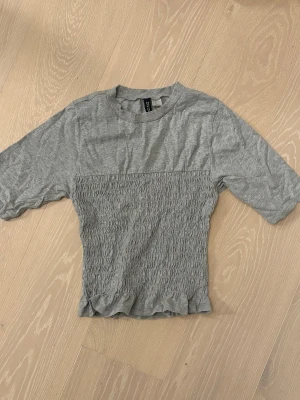 Grå ribbad topp  - Grå topp från DIVIDED i storlek XS med ribbad rund hals och korta ärmar. Nedre delen av toppen har ett smockat, rynkat parti som ger en cool textur och lite volang i nederkant. Perfekt för dig som gillar enkel men snygg stil.