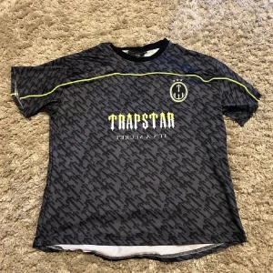 Trapstar svart fotbollströja S - Snygg svart fotbollströja från Trapstar i storlek S med grått camouflage-mönster och gula detaljer. Tryck med Trapstar-logga och nummer 22 på ryggen. Rund hals och korta ärmar. Tillverkad i lätt och ventilerande polyester, perfekt för match eller träning.