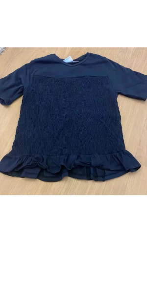 Mörkblå blus med volangkant - Snygg mörkblå blus med korta ärmar och en smockad front. Blusen har en volangkant längst ner som ger en extra touch. Materialet känns lätt och mjukt, perfekt för dig som gillar stilrena detaljer och vill ha något som sticker ut lite.