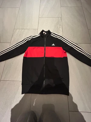 Svart och röd Adidas tränings tröja. 12/13 år. Pojke - Säljer en snygg Adidas vindjacka i svart med en bred röd panel över bröstet och ryggen. Jackan har vita ränder längs ärmarna, hel dragkedja framtill och klassisk Adidas-logga på bröstet. Perfekt för dig som gillar sportig stil.