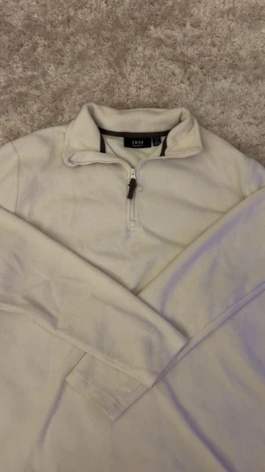 Vit fleece-tröja med half zip från SMOG - Mjuk och skön vit fleece-tröja från SMOG i storlek S. Tröjan har en hög krage och praktisk half zip framtill med en snygg dragkedjedetalj. Regular fit och långärmad, perfekt för kyliga dagar. Enkel och stilren design. Funkar också för olika stilar
