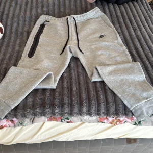 Grå mjukisbyxor från Nike - Grå mjukisbyxor från Nike med svart logga på vänster ben. Byxorna har svarta snören i midjan, ribbade muddar vid bensluten och en dragkedjeförsedd ficka på höger sida. Tillverkade i mjukt bomullsmaterial för maximal komfort.