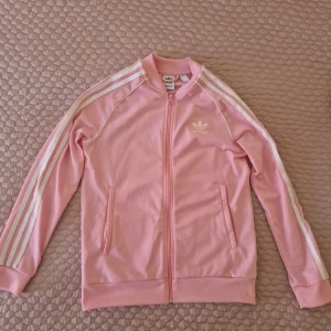 Rosa Adidas ziptröja med vita ränder - Säljer en snygg rosa Adidas ziptröja med klassiska vita ränder längs ärmarna och Adidas-logga på bröstet. Tröjan har dragkedja framtill, ribbade muddar och fickor med dragkedja. ( Små fläckar,se bild) men inget som syns "sticker ut" 