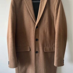 Klassisk brun kappa från New Look Men - Stilren brun kappa från New Look Men i klassisk modell med tre knappar framtill och två fickor med lock. Jackan har snygg krage och är helfodrad, perfekt för höst och vinter. Materialet är mjukt och känns lyxigt.