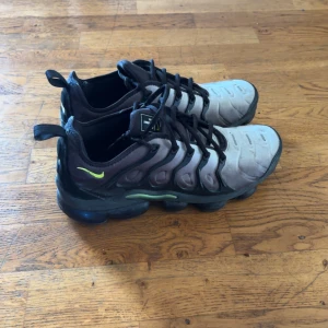 Nike Air Vapormax Plus sneakers - Nike Air Vapormax Plus sneakers i svart och grått med neongröna detaljer. Ovandel i syntet med coola vågformade linjer och svart snörning. Sulan är rejäl och har den ikoniska Vapormax-bubblan för extra komfort och stil. Perfekta för dig som gillar streetwear och unika sneakers. Ser lite använda ut annars bra