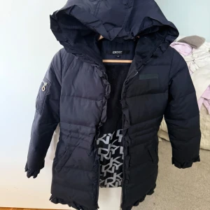Mörkblå pufferjacka från DKNY med huva - Mörkblå pufferjacka från DKNY i storlek M för barn! ca 6-9 År. Jackan har huva, dragkedja, snygga volangdetaljer längs öppningen och är helfodrad med DKNY-logga på insidan. Perfekt för kalla dagar och riktigt skön. Dragkedjan bör lagas och den är märkt med tuschpenna i innerfodret därav priset prutat och klart!