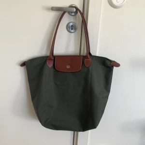 Grön Longchamp axelväska med skinn - Stilren grön axelväska från Longchamp med bruna skinnhandtag och detaljer. Väskan har klassisk form, guldknapp och dragkedja upptill. Perfekt storlek för att få plats med allt du behöver. Materialet är slitstark polyester med snygga kontraster i skinn.