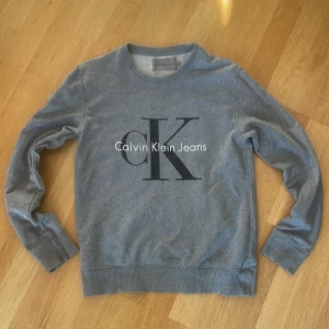 Grå sweatshirt från Calvin Klein Jeans - Snygg grå sweatshirt från Calvin Klein Jeans med stort CK-tryck på bröstet. Tröjan har rund hals, långa ärmar och ribbade muddar vid ärmslut och nederkant. Perfekt för en chill och stilren look.