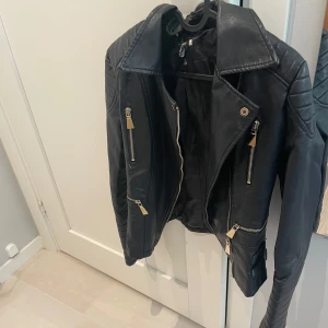 Svart skinnjacka med dragkedjor - Snygg svart skinnjacka med flera silvriga dragkedjor och markerade axlar. Jackan har klassisk krage och quiltade detaljer på axlar och ärmar. Perfekt för dig som vill ha en edgy look.