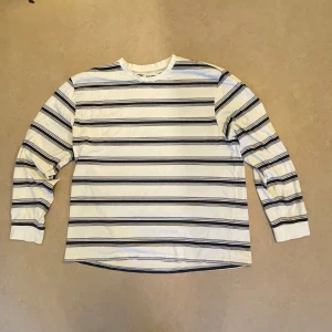 Randig långärmad tröja  - Vit långärmad tröja  med svarta och blå horisontella ränder. Tröjan har rund halsringning och är gjord i mjuk bomull, perfekt för en chill och stilren look.