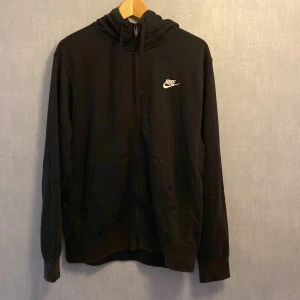 Svart Nike hoodie med dragkedja M - Svart hoodie från Nike i storlek M med vit logga på bröstet. Tröjan har huva med snörning, dragkedja framtill och två stora fickor. Perfekt för chill dagar eller när du vill ha en sportig look.