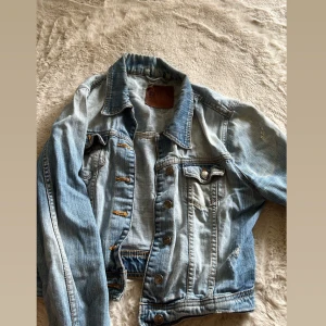 Ljusblå jeansjacka med knappar - Snygg ljusblå jeansjacka i klassisk modell med metallknappar framtill och på bröstfickan. Jackan har en krage, synliga sömmar och slitna detaljer för en cool, avslappnad look. Perfekt att slänga över en hoodie eller t-shirt.