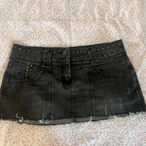 Svart jeanskjol med nitar - Cool svart jeanskjol med slitna detaljer och rå kant nertill. Kjolen har silverfärgade nitar längs midjan och fickorna, vilket ger en edgy vibe. Klassisk femficksmodell och knappstängning framtill. Perfekt för dig som gillar en tuffare stil.