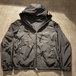 Grå vindjacka från Moncler med huva - Grå vindjacka från Moncler med huva och dragsko, hel dragkedja framtill och två fickor med tryckknappar. Jackan har Moncler-logga på ena fickan och diskreta rödvita detaljer på huvan. Perfekt för dig som vill ha en stilren och sportig look. Hör av vid minsta fundering 💫