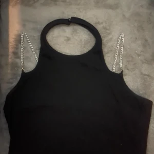 Svart ärmlös topp med kedjedetaljer - Snygg svart ärmlös topp med hög hals och coola silverfärgade kedjor som axelband. Toppen har en stilren och modern look, perfekt för dig som vill sticka ut. Materialet känns mjukt och följsamt mot kroppen.
