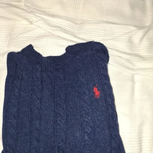 Mörkblå kabelstickad tröja Polo Ralph Lauren - Kabelstickad mörkblå tröja från Polo Ralph Lauren med klassisk röd broderad logga på bröstet. Tröjan har rund halsringning och långa ärmar, perfekt för lager-på-lager. Tillverkad i mjukt stickat material som håller dig varm och snygg. Priset kan diskuteras 