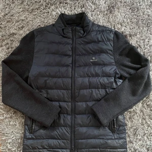 pufferjacka från GANT - Säljer en pufferjacka från GANT med quiltad framsida och baksida i glansigt material. Ärmarna är i mjuk, mörkgrå stickad kvalitet. Jackan har dragkedja framtill och två fickor med dragkedja. Perfekt för kyliga dagar.
