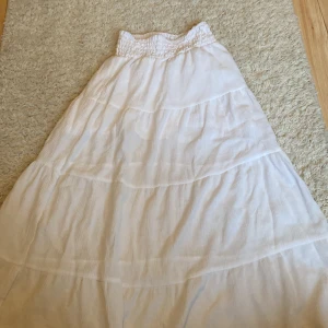 Vit långkjol från H&M med volanger - Superfin vit långkjol från H&M med smock i midjan och flera volanglager. Kjolen är luftig och har en lätt, flowy känsla som passar perfekt till varma dagar. Tillverkad i viskosblandning för extra komfort.