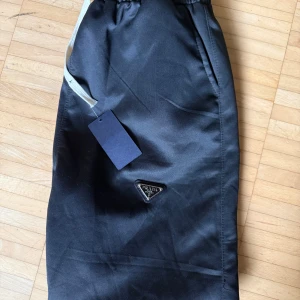 Svarta Prada byxor med logga - Svarta byxor från Prada med elastisk midja och dragsko. Byxorna har en stilren look med diskret Prada-logga framtill och sidfickor. Materialet är glansigt och känns lyxigt, perfekt för dig som gillar exklusiva streetwear-vibbar.