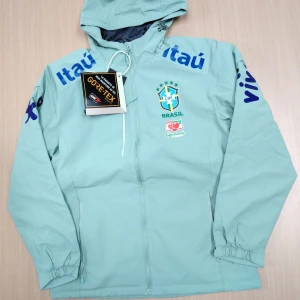 Nike Brazil vindjacka LIGHT PISTACHIO - Säljer en sjukt snygg ljusblå vindjacka från Nike med Brasil-logga och sponsormärken. Jackan har huva, dragkedja framtill och elastiska muddar. Materialet är lätt och vattentätt Gore-Tex, perfekt för blåsiga dagar. Stora tryck på ärmarna och bröstet ger en sportig vibe.