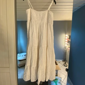 Vit kort kläning med smock - Säljer en vit kort kläning med smock upptill och tunna axelband. Klänningen har en luftig och böljande form med lätt rynkad struktur i tyget, perfekt för varma dagar. Enkel och stilren design som ger en fräsch vibe❤️❤️