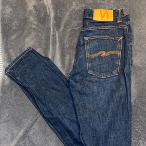 Nudie Jeans Grim Tim Dry Dirt Organic 28/32 - Säljer ett par mörkblå Nudie Jeans Grim Tim Dry Dirt Organic i storlek 28/32. Klassisk femficksmodell med orange sömmar och Nudie-logga på bakfickan. Tillverkade i ekologisk bomull för en schysst känsla och look.