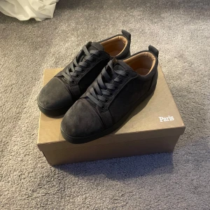 Christian Louboutin gråa utan spikar  - Snygga gråa sneakers från Christian Louboutin i mocka med klassisk röd sula. Skorna har rund tå, svarta skosnören och beige innersida i skinn. Perfekta för dig som vill ha en lyxig och stilren look med en ikonisk touch.