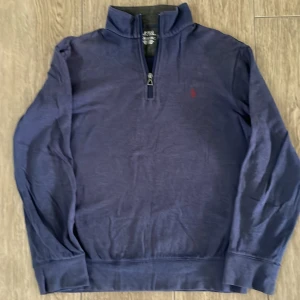 Blå half zip tröja från Ralph Lauren - Mörkblå långärmad tröja från Ralph Lauren med röd broderad logga på bröstet. Tröjan har en half zip-dragkedja vid halsen och är tillverkad i mjuk bomull. Perfekt för lager-på-lager