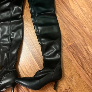 Svarta overknee boots med klack, stl 39 - Säljer ett par svarta overknee boots i storlek 39. Skorna har spetsig tå, smal klack och dragkedja på insidan. Tillverkade i syntetmaterial med en stilren look som passar till många outfits. Perfekta för dig som vill ha ett par höga boots med attityd.