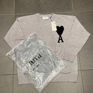 Grå stickad tröja från Ami Paris - Snygg ljusgrå stickad tröja från Ami Paris med svart hjärta och A-logga på bröstet. Tröjan har rund hals, ribbade muddar och är långärmad. Perfekt för dig som gillar stilrena detaljer och vill ha något som sticker ut lite extra.