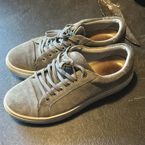 Grå mockasneakers från Vagabond - Snygga grå sneakers från Vagabond i mocka med klassisk rund tå och platt vit sula. Skorna har snörning med matchande grå skosnören och diskreta sömdetaljer. Perfekta för en clean och stilren look.