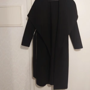 Svart lång kappa med bälte - Stilren svart kappa med lång passform och bred krage. Kappan har ett bälte i midjan för att kunna knytas och långa ärmar. Perfekt för dig som gillar en klassisk look med modern touch.Lite nopprigt under armhål kolla bilden.