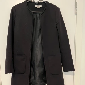 Svart rock med fickor från Filippa K - Stilren svart rock från Filippa K med rundad halsringning och två stora fickor framtill. Jackan är helfodrad och har en rak, klassisk passform som ger en clean look. Perfekt för dig som gillar minimalistisk stil.