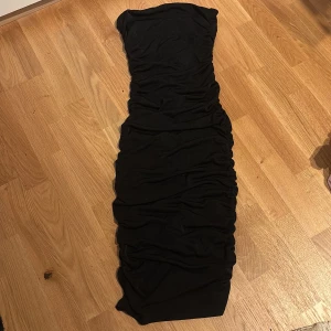 Svart glittrig långklänning - Supersnygg svart långklänning från Amisu med glittrigt tyg och figurnära passform. Klänningen är axelbandslös och har rynkade detaljer längs hela längden. Perfekt för dig som vill sticka ut med stil och passar för alla tillfällen🖤✨