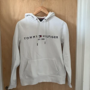 Vit Tommy Hilfiger hoodie - Vit hoodie från Tommy Hilfiger med broderad logga och text på bröstet. Klassisk känguruficka, snörning i huvan och liten flagga på ärmen. Mjuk insida och normal passform, perfekt för chill dagar.
