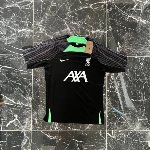 Liverpool FC svart tränings-tshirt Nike - Liverpool FC tränings-tshirt från Nike i svart med gråmönstrade ärmar och gröna detaljer vid hals och sidor. Stor vit AXA-logga på bröstet och klubbmärke. Tillverkad i lätt och ventilerande funktionsmaterial, perfekt för fotbollsträning.