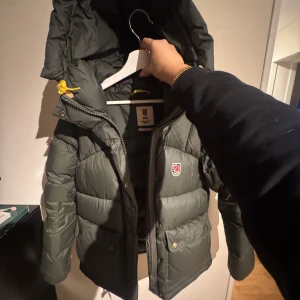 Grön Fjällräven Expedition dunjacka - Säljer en grön Fjällräven Expedition Series dunjacka med huva och gula snören. Jackan har två stora fickor med knappar, dragkedja och Fjällräven-logga på bröstet. Puffad design och långärmad, perfekt för kalla dagar. Riktigt snygg och klassisk look.