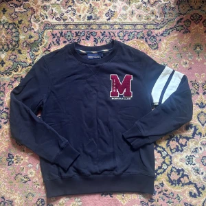 Mörkblå sweatshirt Morris R. Club S - Mörkblå sweatshirt från Morris R. Club med stor röd M-applikation och broderad text på bröstet. Vit och blå randig detalj på ena ärmen. Klassisk rund hals och ribbade muddar. Tillverkad i mjuk bomull, perfekt för chill dagar.